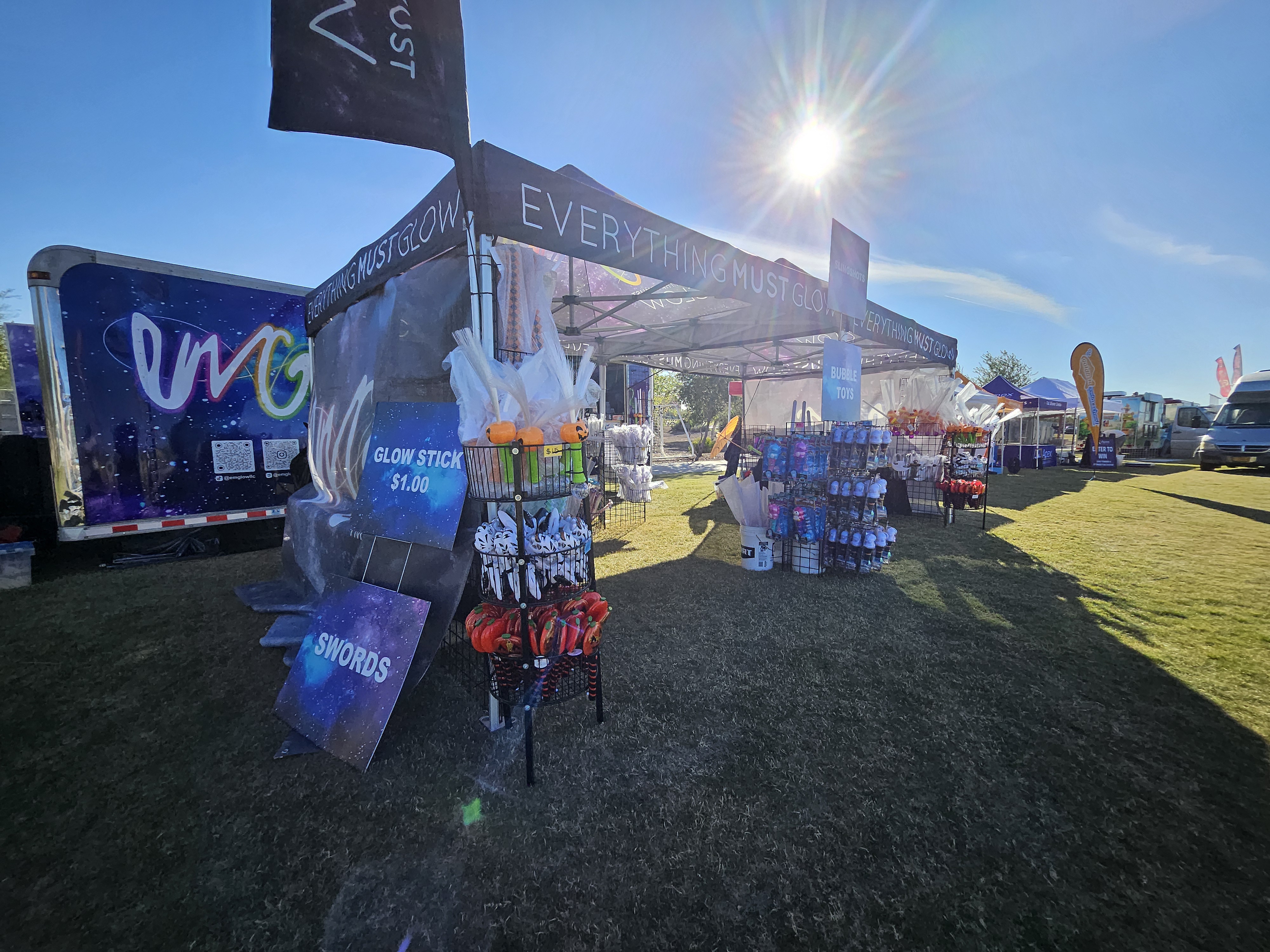 Daytime booth setup with glow stick signs, swords, and the branded trailer in the sun