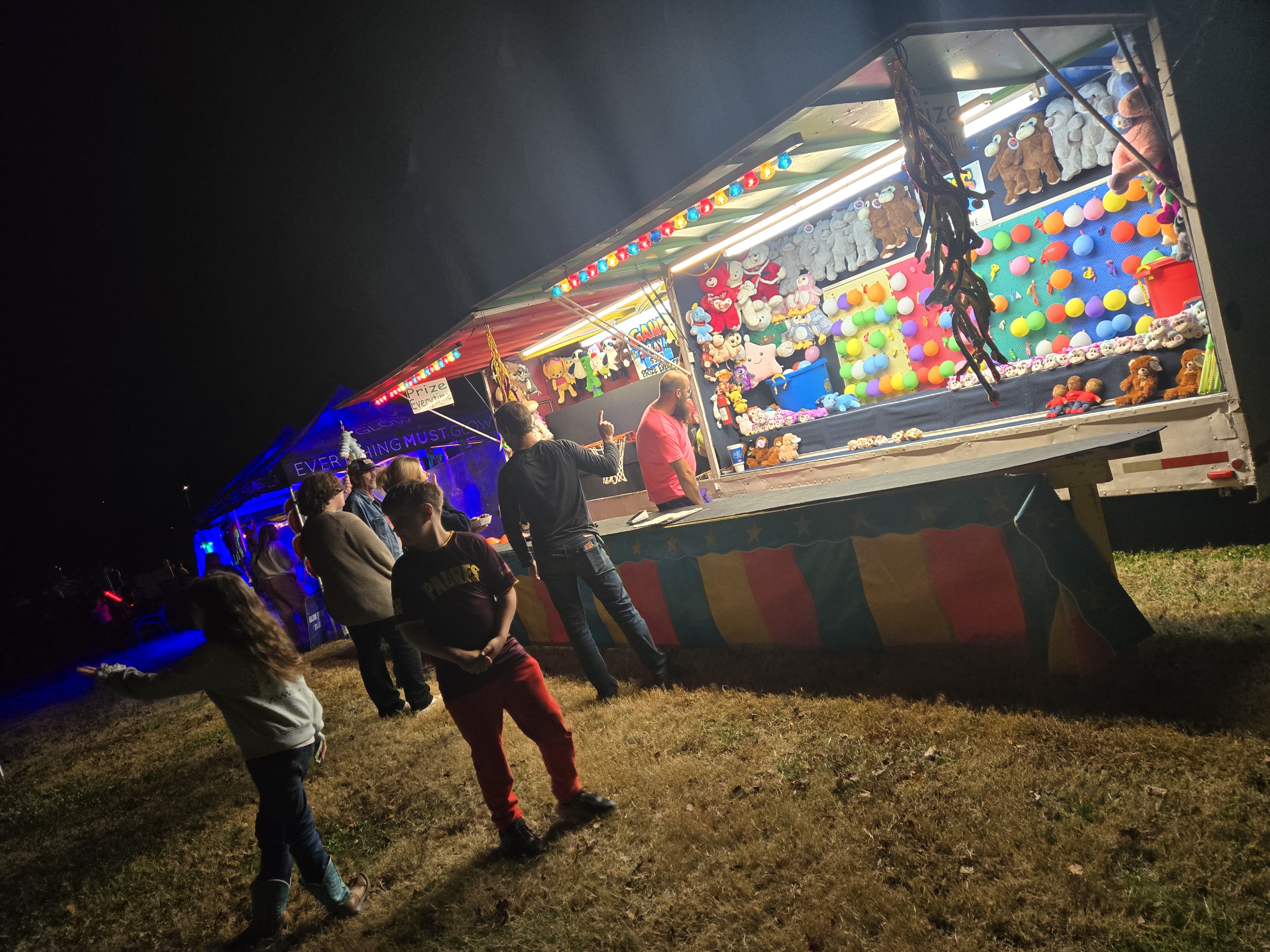Carnival balloon dart game lit up at night with our booth glowing in the background