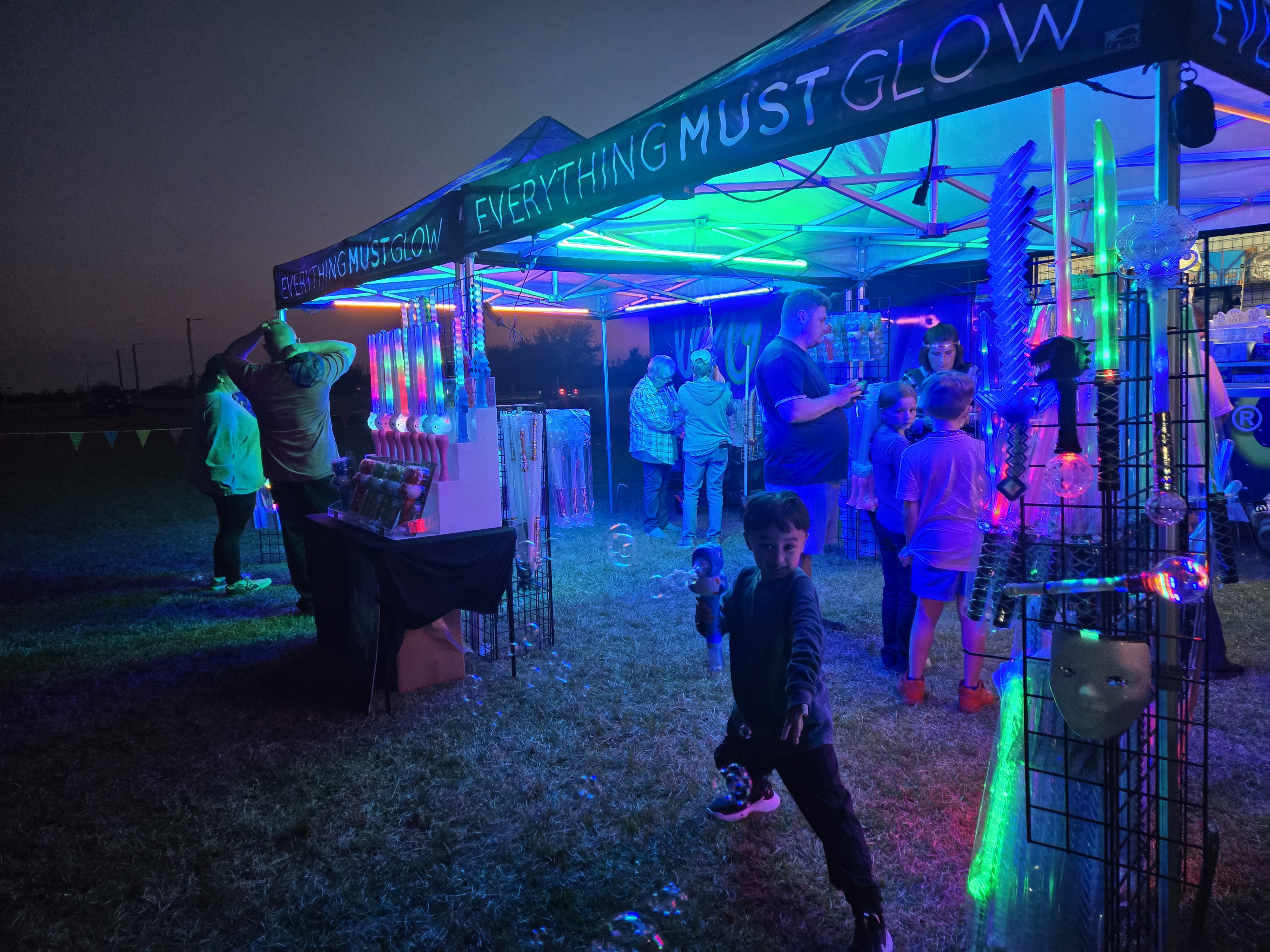 The Everything Must Glow booth lit up with UV lights, kids blowing bubbles, LED swords everywhere